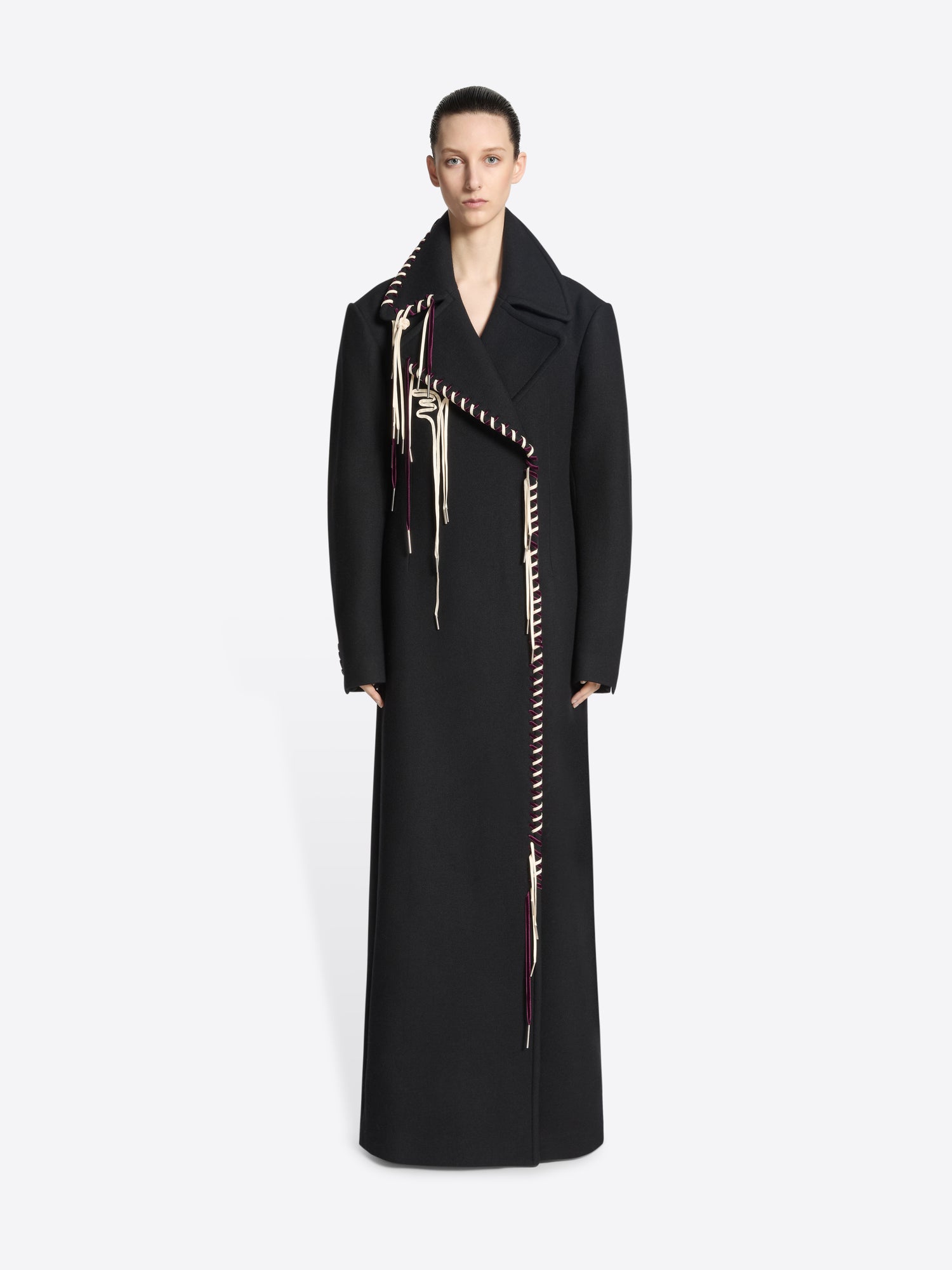 Person wearing a long belted black coat with white and burgundy detailing on white background