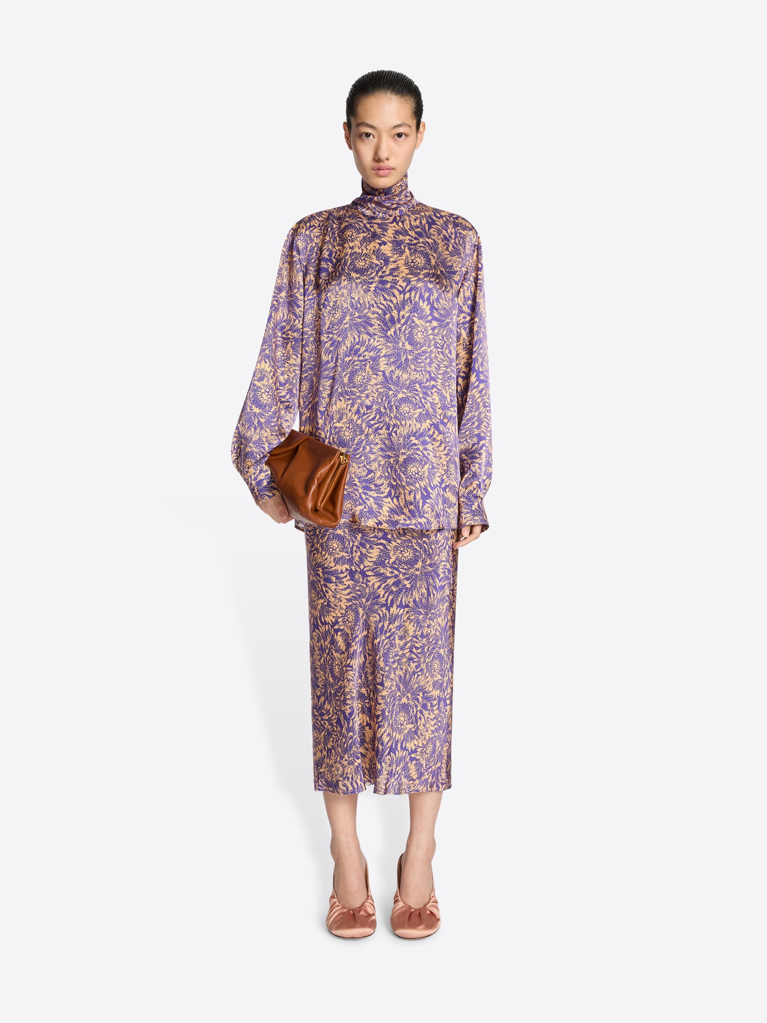 Woman wearing a purple floral dress holding a brown clutch against a white background