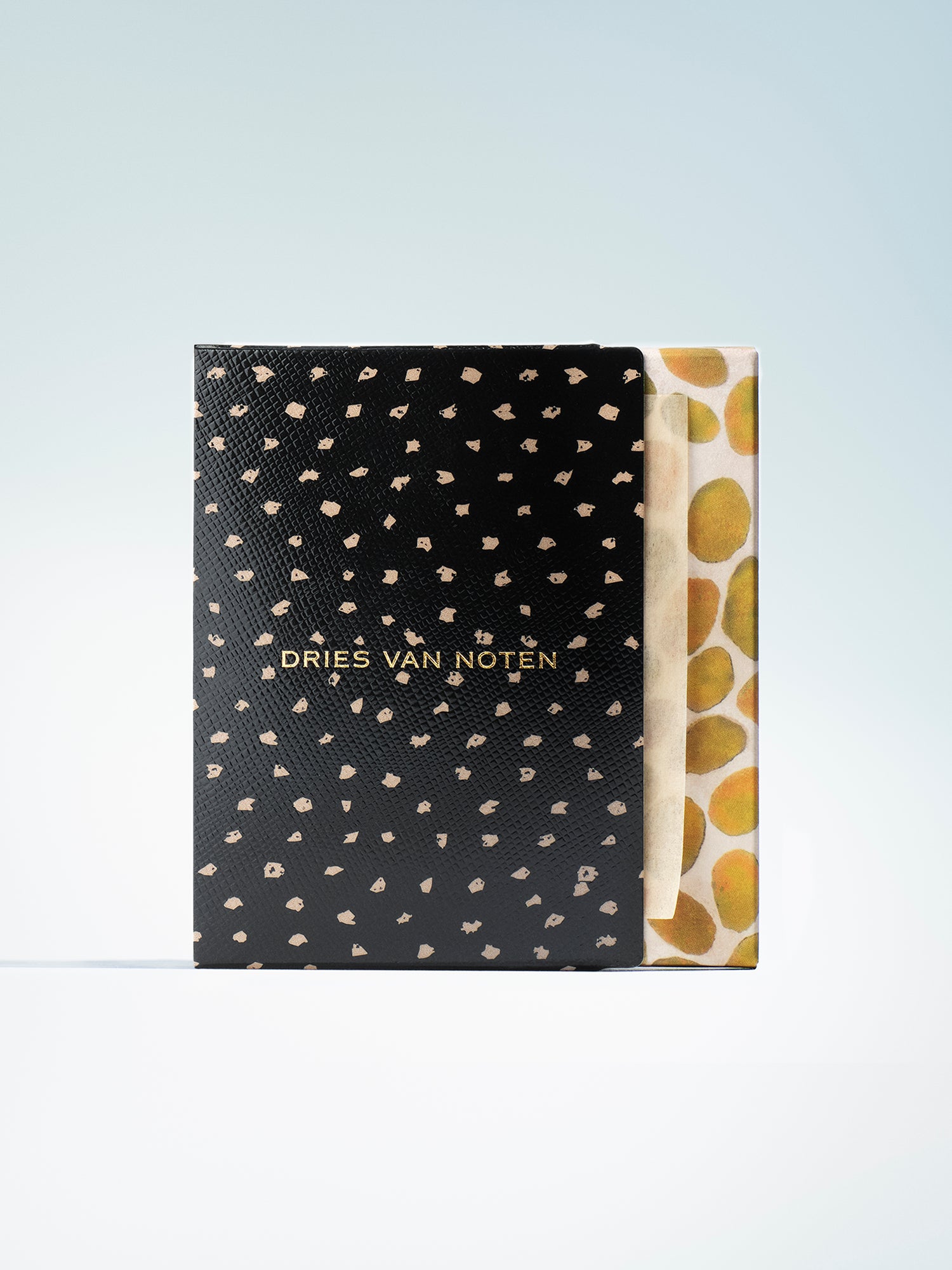 A Dries Van Noten lip blotting papers set standing upright, showing a black textured cover with scattered small gold spots and gold lettering, next to a cream paper sleeve with large gold circular motifs, on a light blue gradient background.