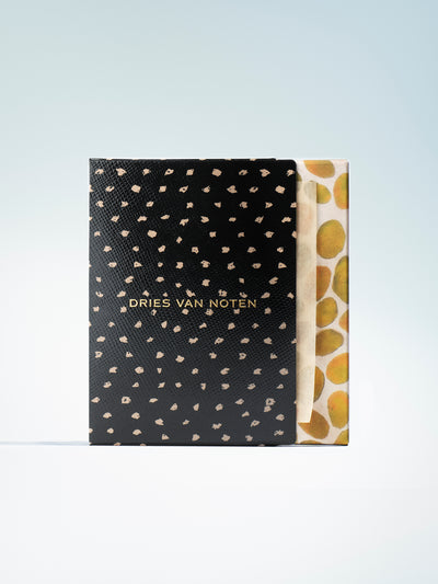 A Dries Van Noten lip blotting papers set standing upright, showing a black textured cover with scattered small gold spots and gold lettering, next to a cream paper sleeve with large gold circular motifs, on a light blue gradient background.
