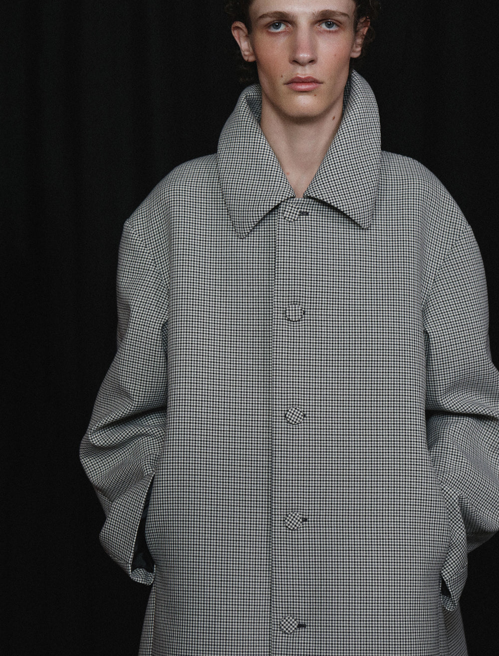 Model wearing a gray checkered coat against a black curtained background