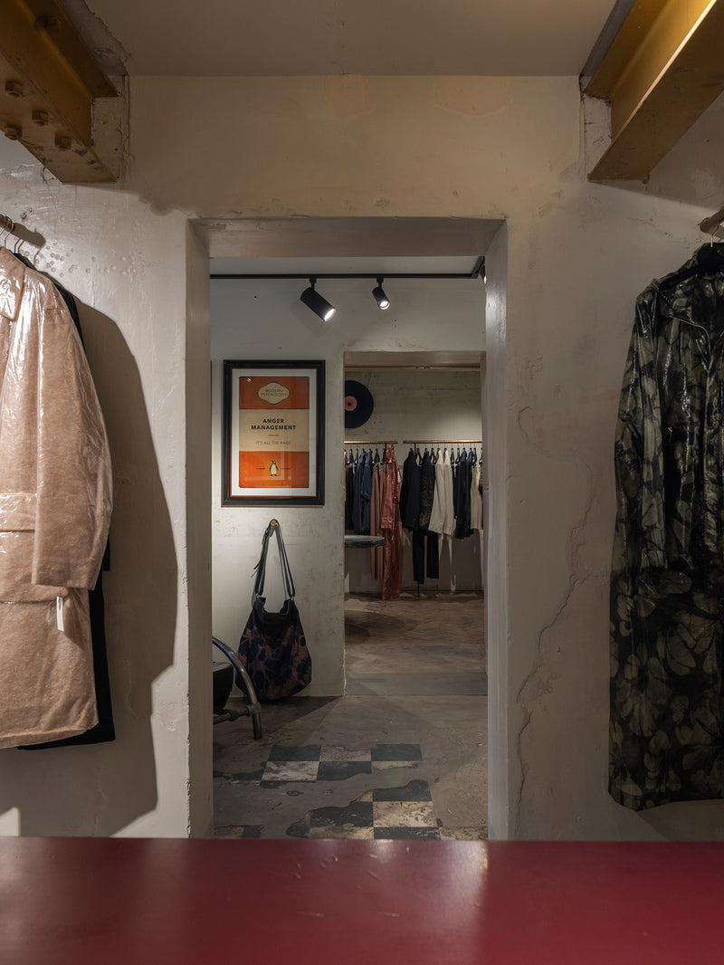 A view from inside a clothing boutique looking through a doorway, with garments hanging on the left and right walls, a framed poster on the back wall, and a red tabletop in the foreground.