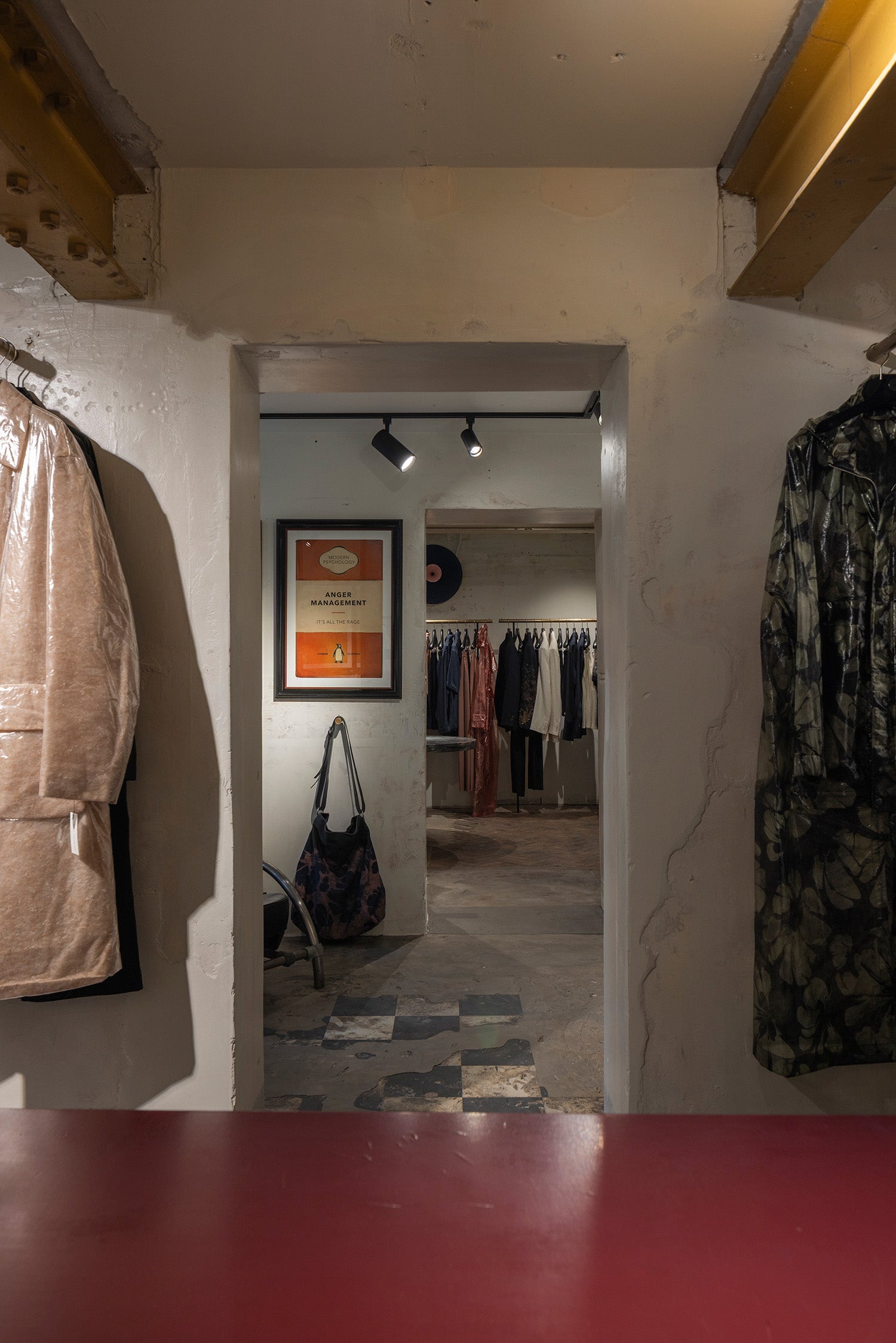 A view from inside a clothing boutique looking through a doorway, with garments hanging on the left and right walls, a framed poster on the back wall, and a red tabletop in the foreground.