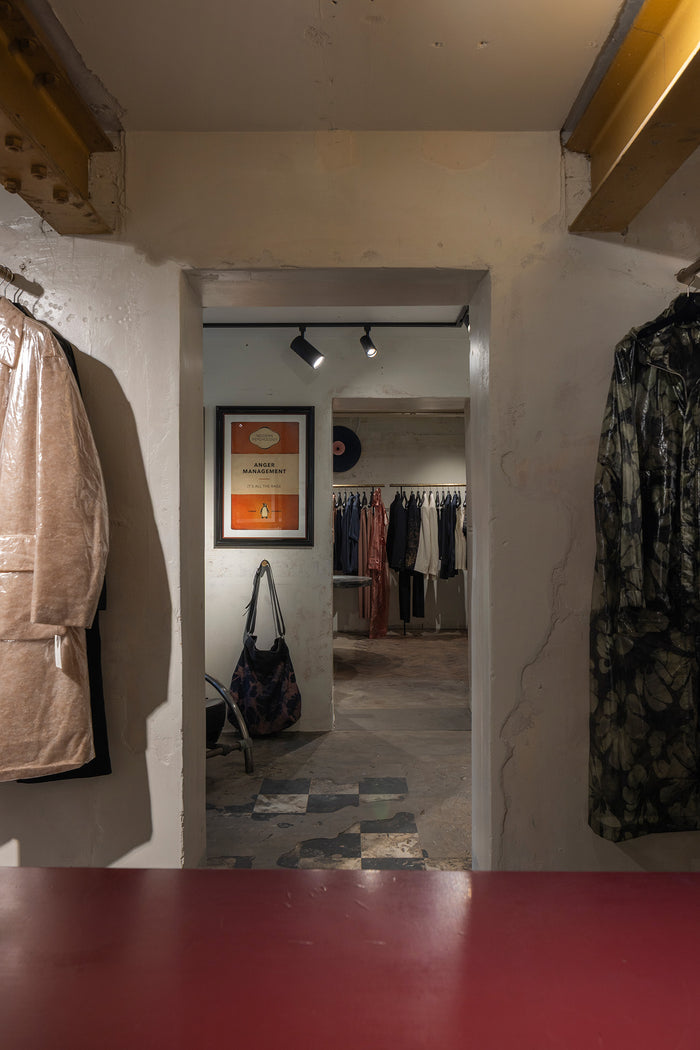 A view from inside a clothing boutique looking through a doorway, with garments hanging on the left and right walls, a framed poster on the back wall, and a red tabletop in the foreground.