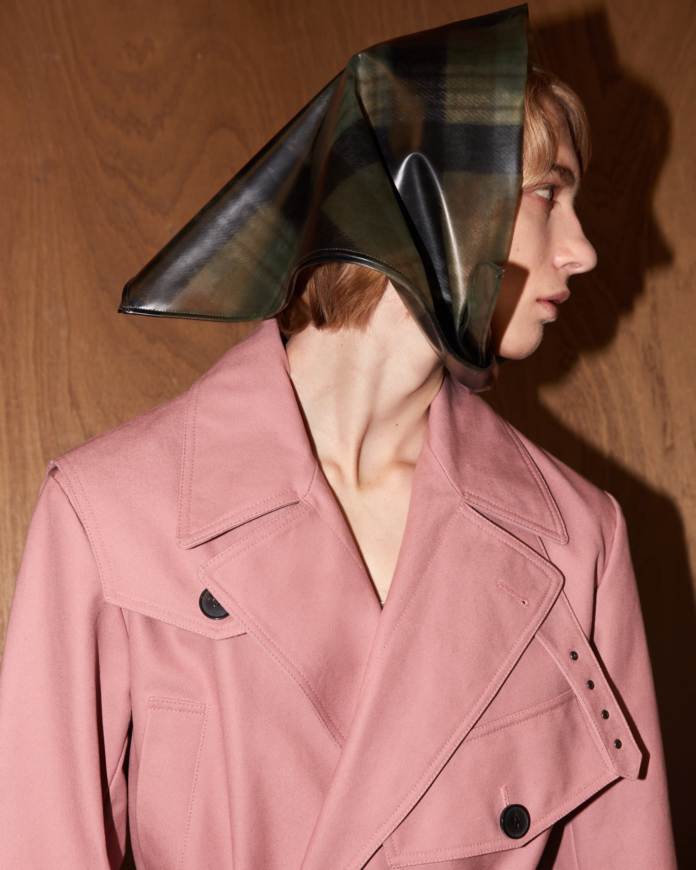 Person wearing a pink coat with a unique bandana headpiece against a wooden background