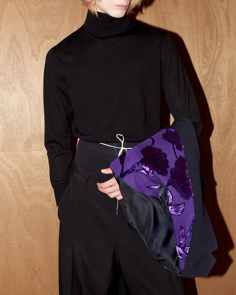 Person wearing a black turtleneck and holding a purple floral blazer against a wooden background.
