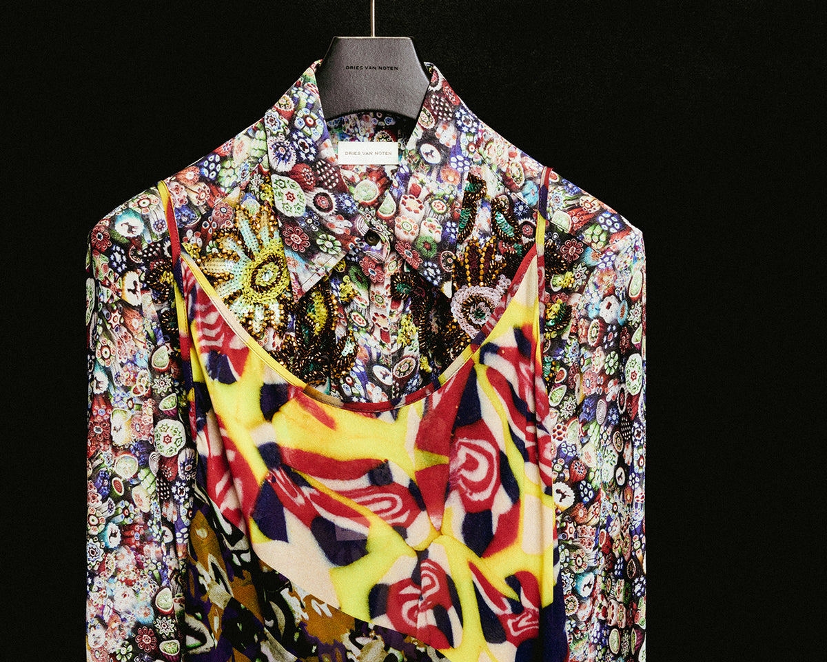 A colorful silk shirt and dress with a highly detailed, multicolored floral and abstract pattern, draped over a black hanger against a solid black background.