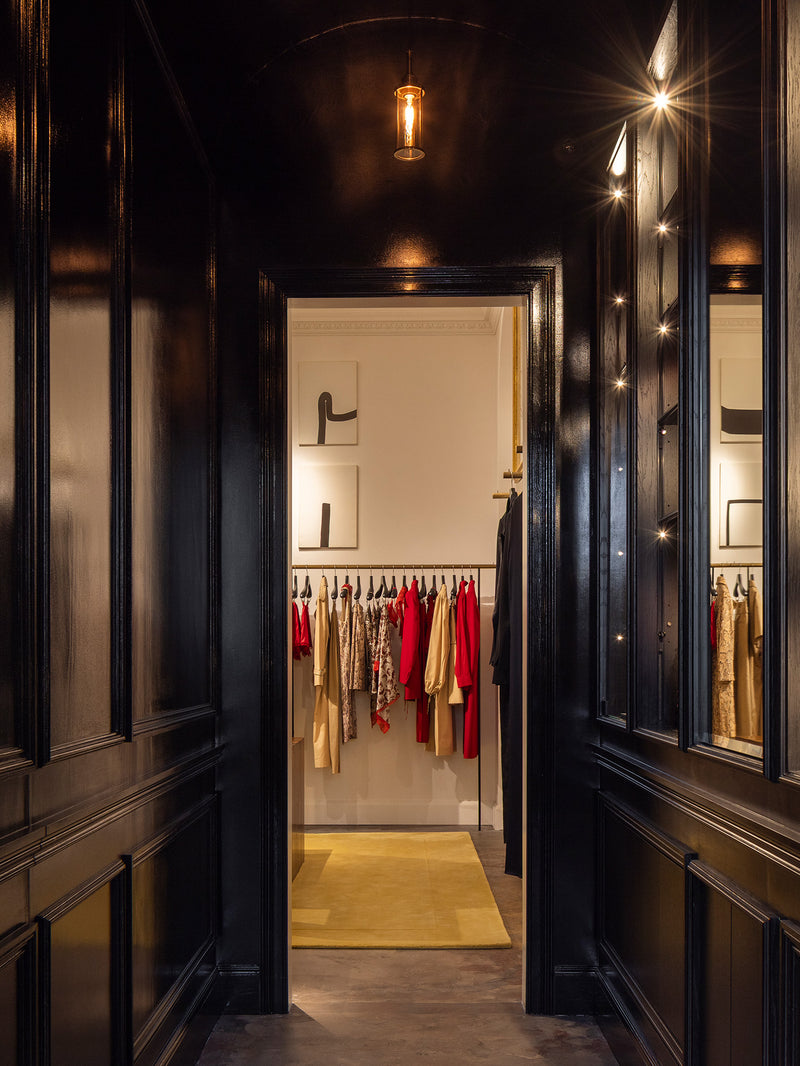 A dark, elegant interior hallway with glossy black wood paneling and mirrors, looking through an open doorway into a rom filled with hanging garments.
