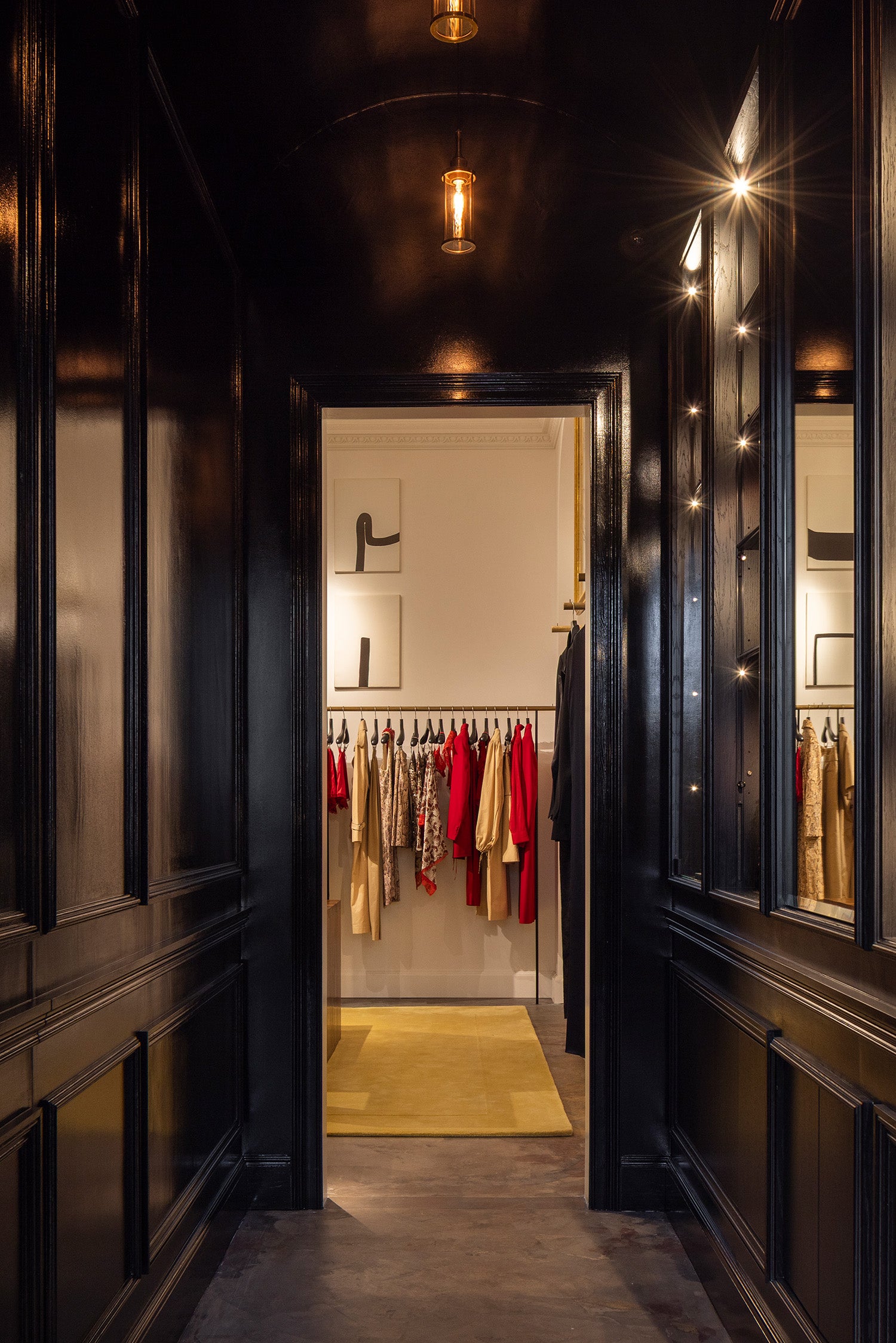 A dark, elegant interior hallway with glossy black wood paneling and mirrors, looking through an open doorway into a rom filled with hanging garments.