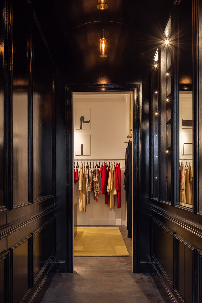 A dark, elegant interior hallway with glossy black wood paneling and mirrors, looking through an open doorway into a rom filled with hanging garments.