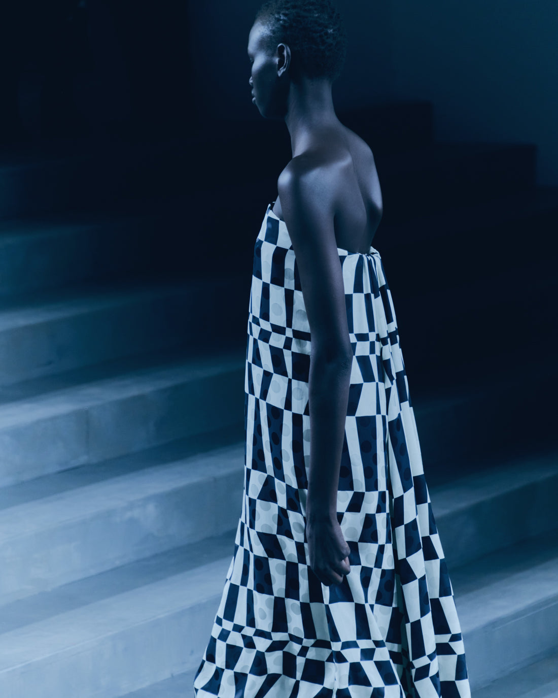 Woman wearing a black and white checkered dress in a dimly lit setting with a staircase in the background