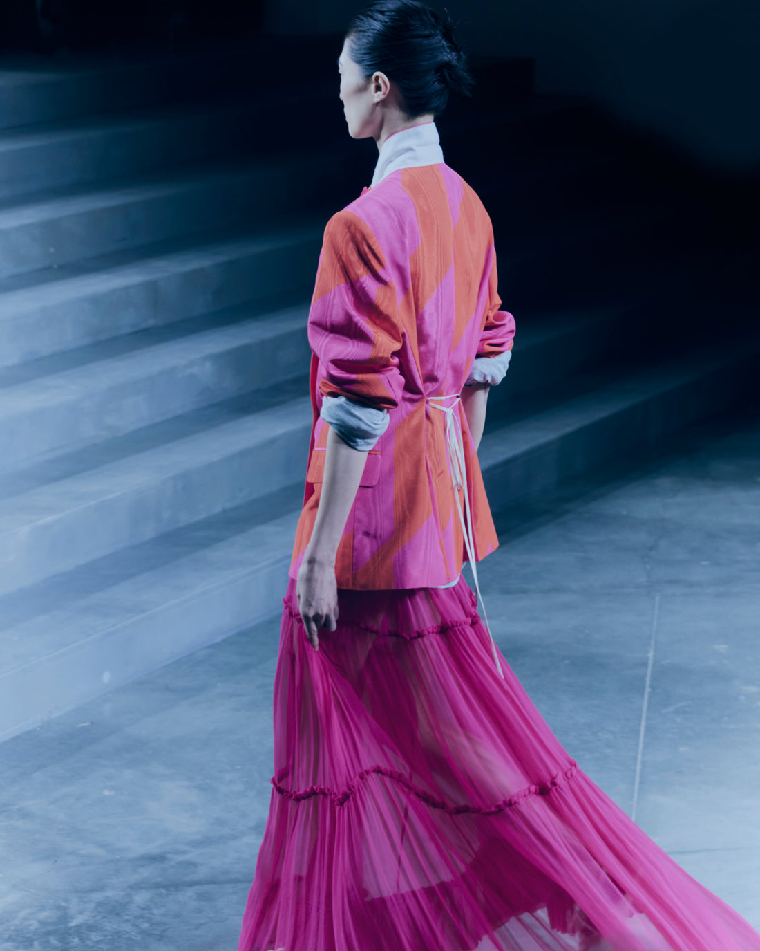 Model wearing a vibrant outfit with a pink and orange blazer and a long pink skirt on a staircase.