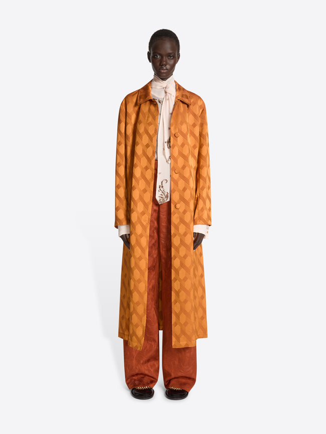 Person wearing a white top, dark orange pants and an orange jacquard mac with chain motif on a white background