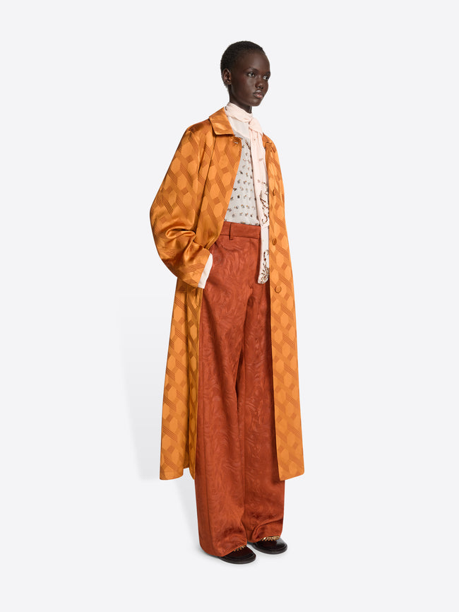 Person wearing a white top, dark orange pants and an orange jacquard mac with chain motif on a white background