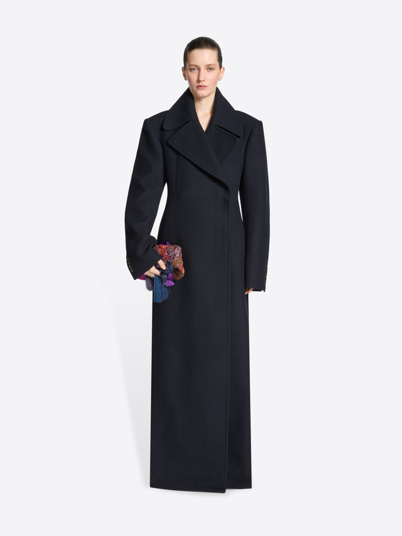 Person wearing a navy coat with a belt, carrying an embellished handbag on a white background