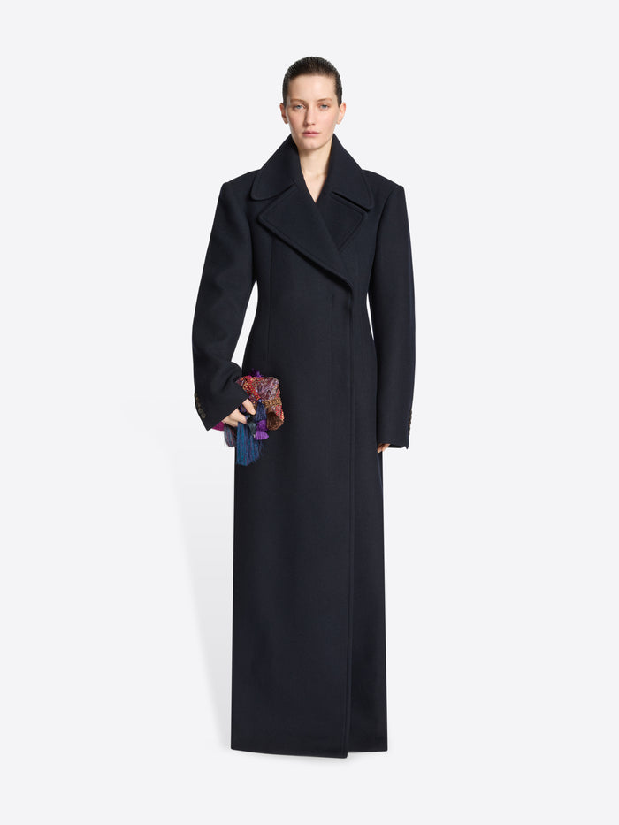 Person wearing a navy coat with a belt, carrying an embellished handbag on a white background