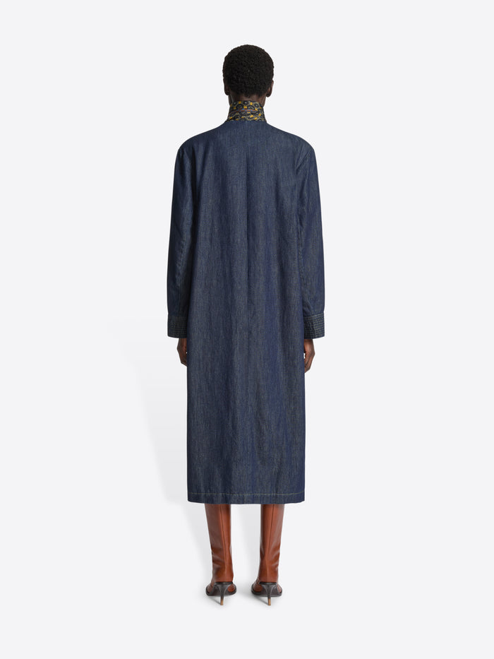 Backside of a person wearing a long collarless denim coat on a white background