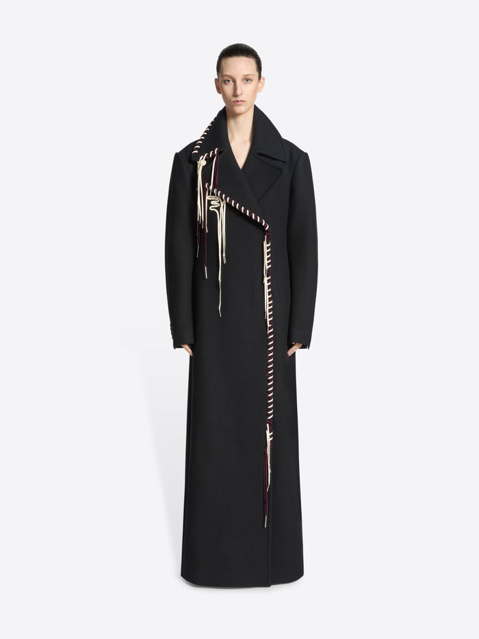 Person wearing a long belted black coat with white and burgundy detailing on white background 