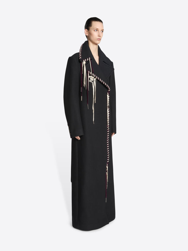 Person wearing a long belted black coat with white and burgundy detailing on white background 