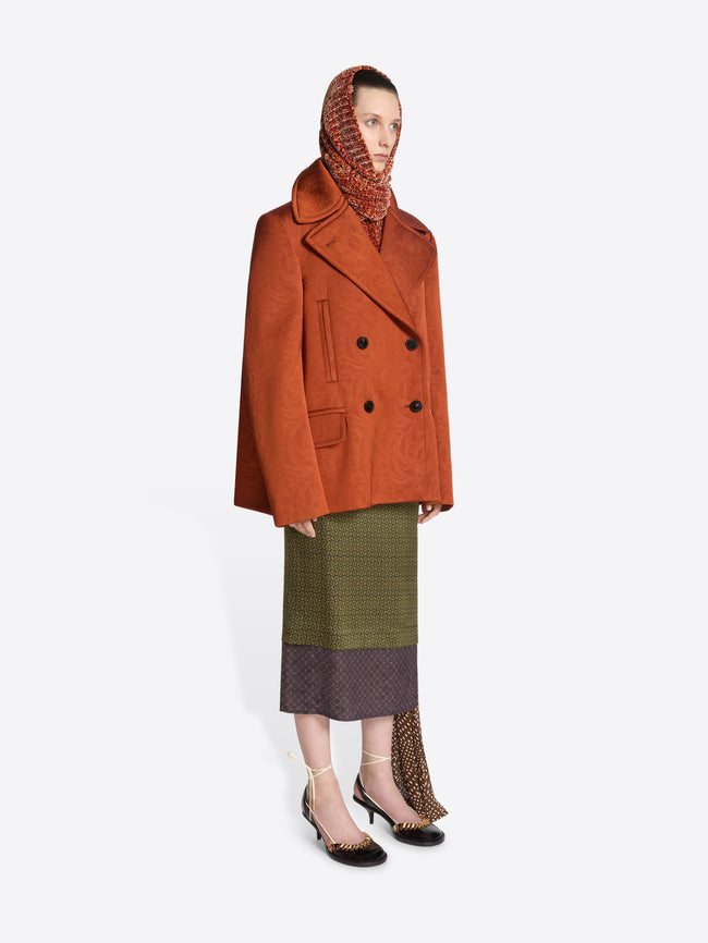 Person wearing a patterned rust coloured coat with brown buttons and a green skirt on a white background