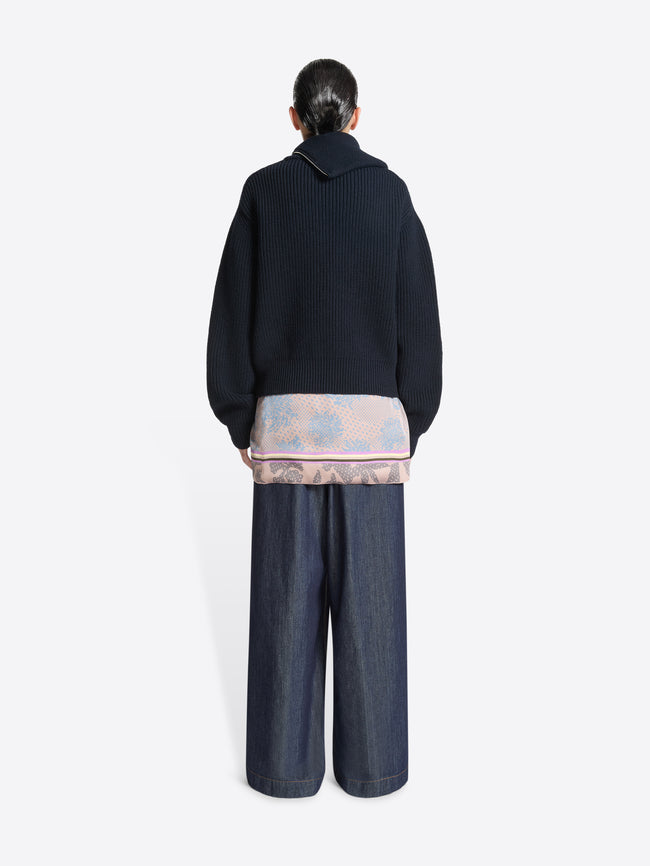 Backside of a person wearing a dark sweater, wide-leg jeans and a light colored, floral patterned, mini wrap skirt on a white background