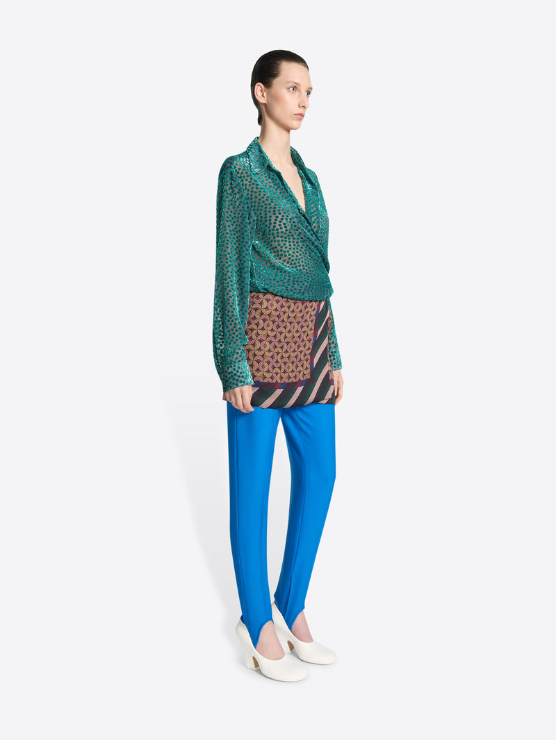 Person wearing blue pants, a wrap skirt and a turquoise patterned and textured shirt with a tie on a white background