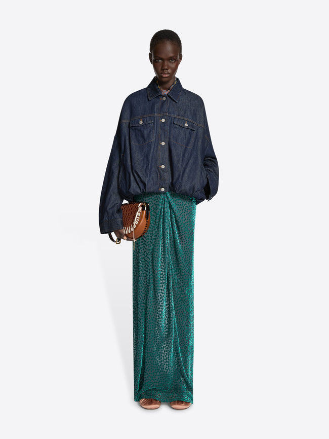 Person wearing a denim top and a textured,  turquoise, floor-length draped skirt while holding a tan colored bag on a white background