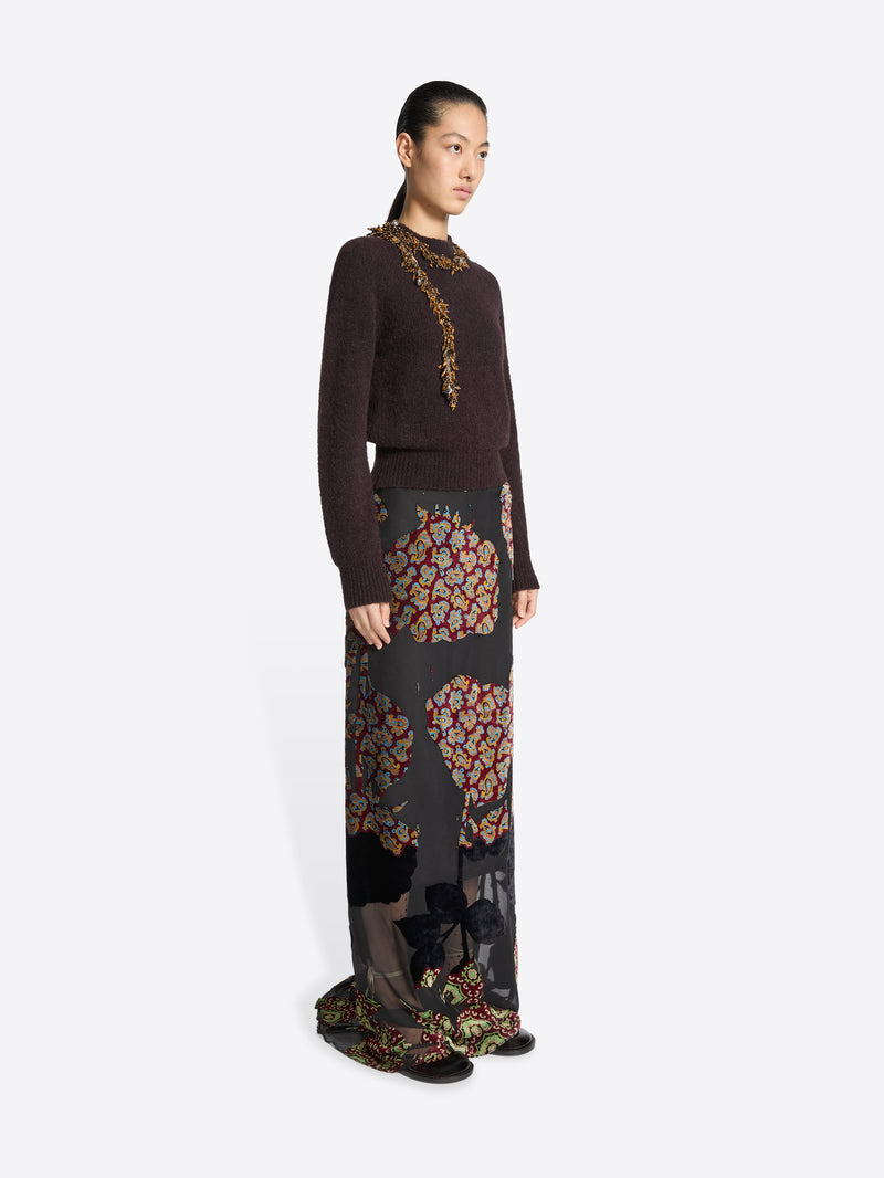 Model wearing a dark brown sweater and long, black, floral patterned skirt on a white background