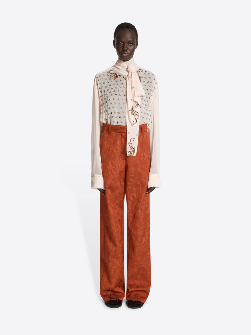 Model wearing an embellished blouse with a scarf and rust coloured textured pants on a white background