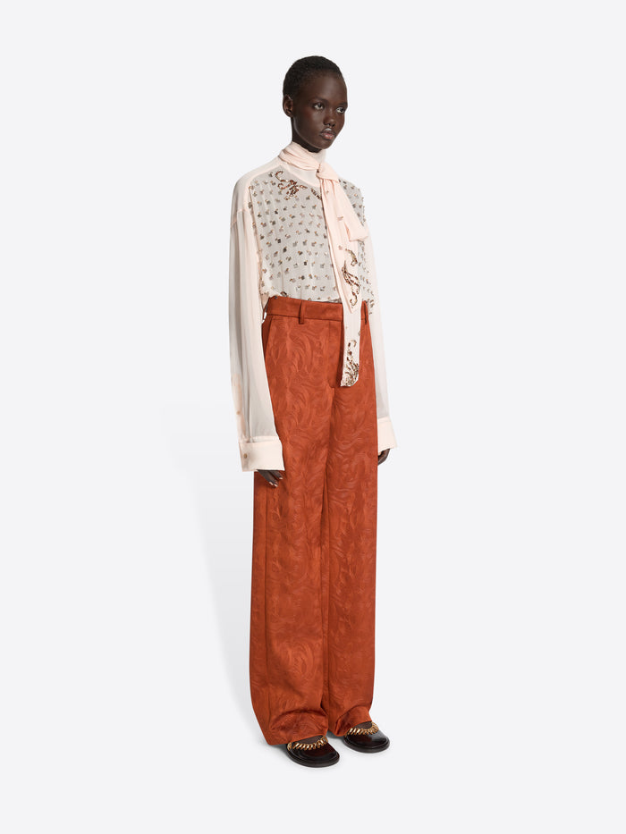 Model wearing an embellished blouse with a scarf and rust coloured textured pants on a white background