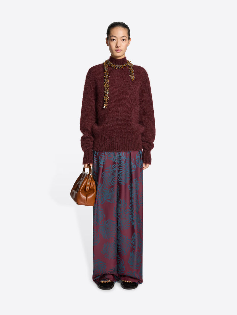 Person wearing a burgundy sweater and patterned pants holding a brown handbag on a white background