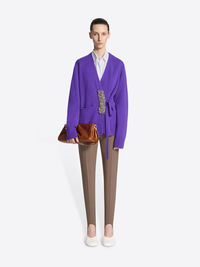 Person wearing a purple sweater with an embellished closure detail, taupe stirrup pants, and white shoes on a white background.