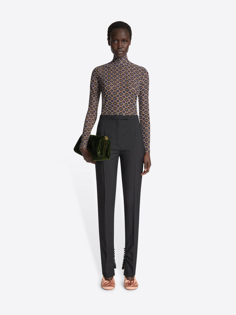 Model wearing a patterned top and black dresspants holding a clutch, on a white background