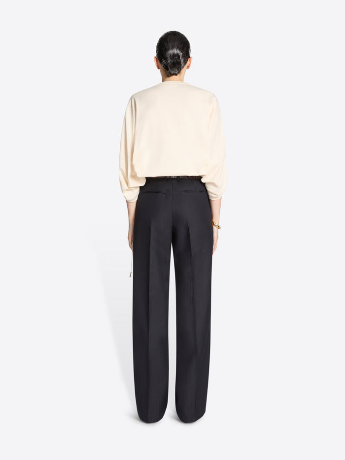 Person wearing a cream coloured sweater and navy pants with white and brown striped side tassels on a white background