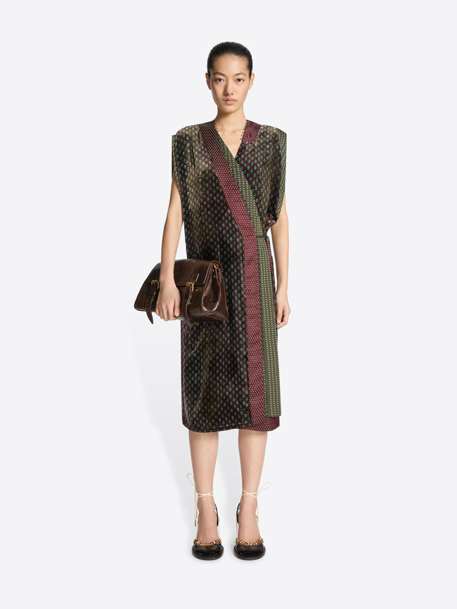 Woman wearing a patterned dress in khaki color mix holding a matching handbag on a white background