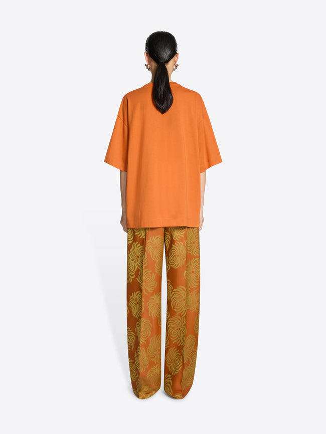 Backside of a person wearing an orange, loose cotton t-shirt and orange pants with a yellow floral pattern on a white background