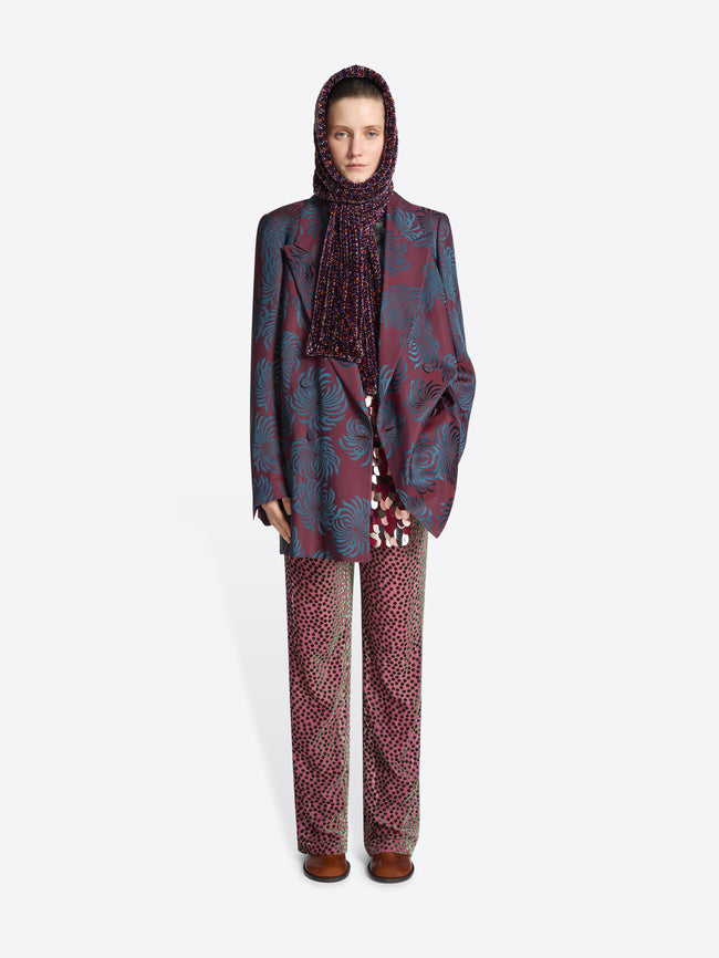 Model wearing a purple colored hooded scarf while wearing a burgundy floral print blazer and pink patterned pants on a white background