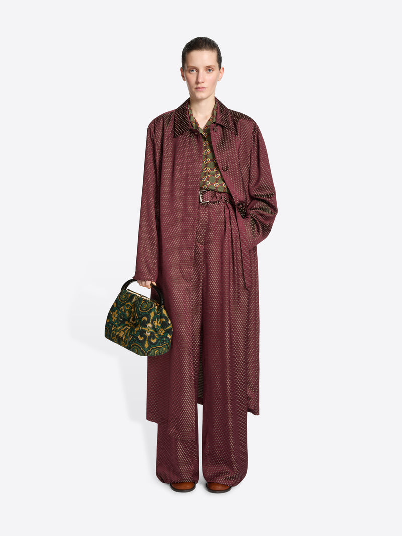 Model wearing a burgundy coloured coat and matching trousers while holding a pleated jacquard bag with green, yellow and black design on a white background