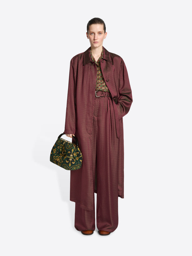 Model wearing a burgundy coloured coat and matching trousers while holding a pleated jacquard bag with green, yellow and black design on a white background