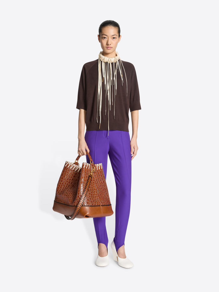 Woman wearing a brown top and purple pants holding a brown handbag on a white background