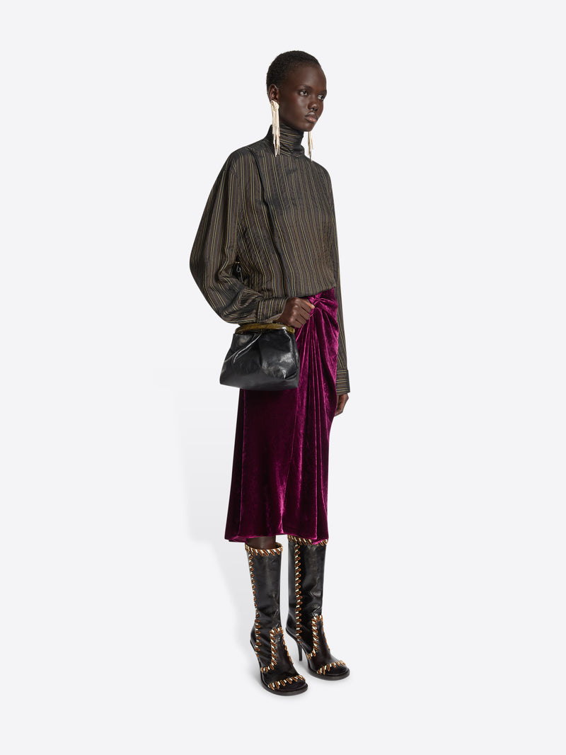 Person wearing a brown striped top and a burgundy velvet skirt, while holding mall pleated leather bag with resin handle on a white background