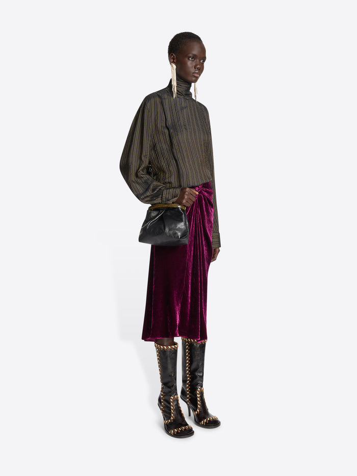Person wearing a brown striped top and a burgundy velvet skirt, while holding mall pleated leather bag with resin handle on a white background