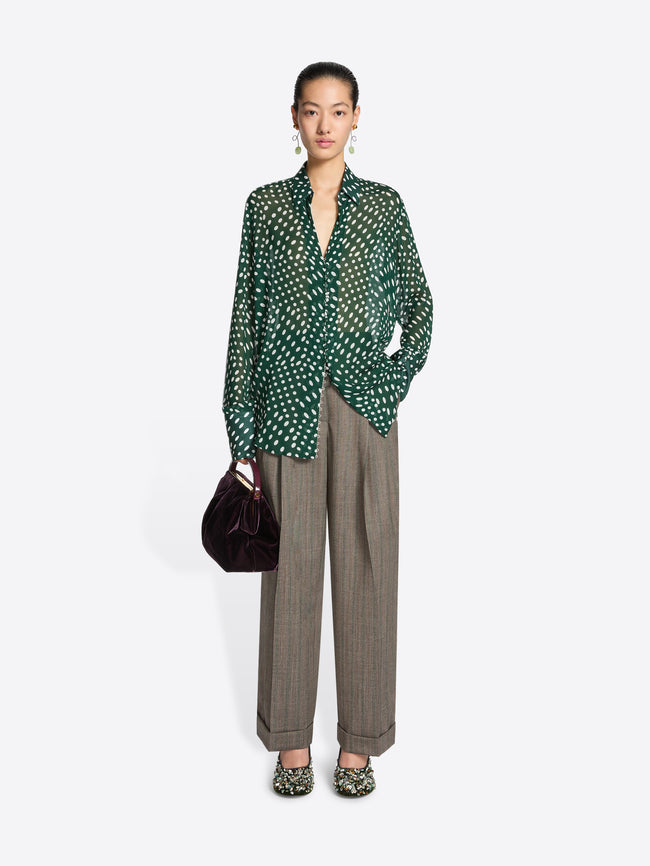 Person wearing a green patterned top, gray pants and a pair of olive colored embellished mules on a white background