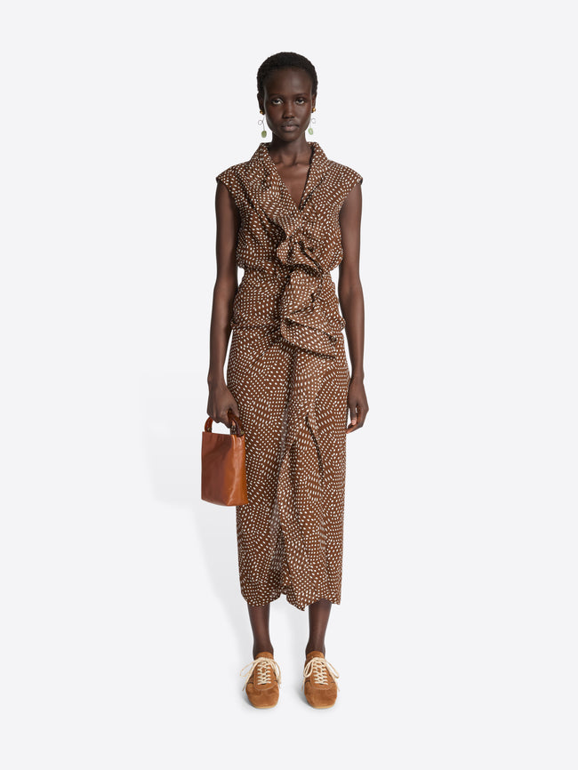 Person wearing a brown patterned dress while holding a tan leather bag and wearing tan suede sneakers on a white background