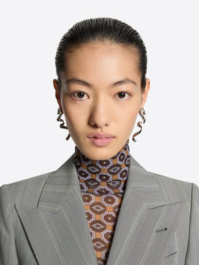 Person wearing a gray suit with a patterned shirt and earrings against a white background