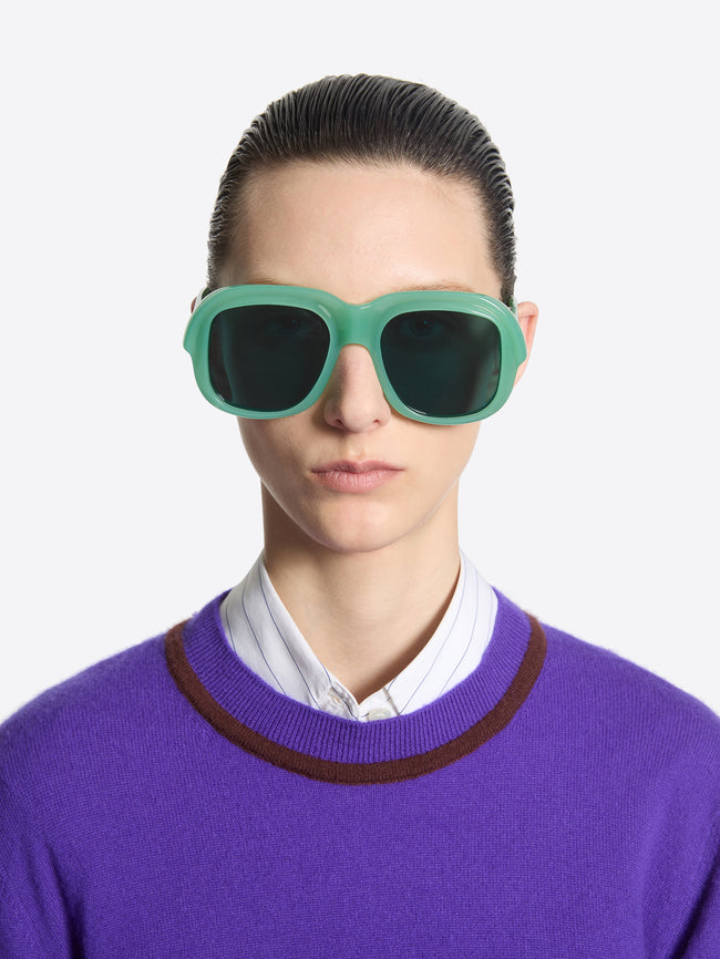 Person wearing green sunglasses and a purple sweater with a white shirt underneath on a light gray background