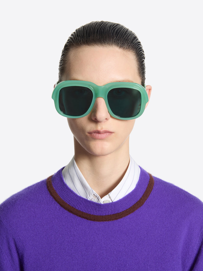 Person wearing green sunglasses and a purple sweater with a white shirt underneath on a light gray background