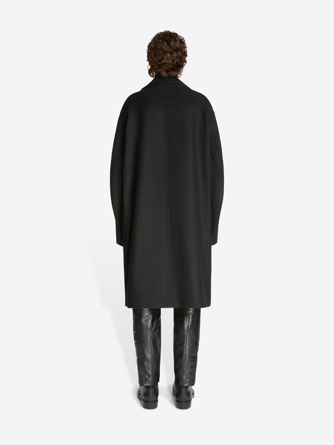 Backside of a model wearing black leather pants, black leather boots and a black, woolen caban coat on a white background