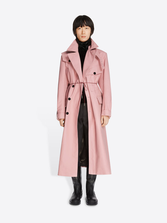 Model wearing a black turtleneck, black pants, black leather boots and a rose colored cotton trench coat on a white background