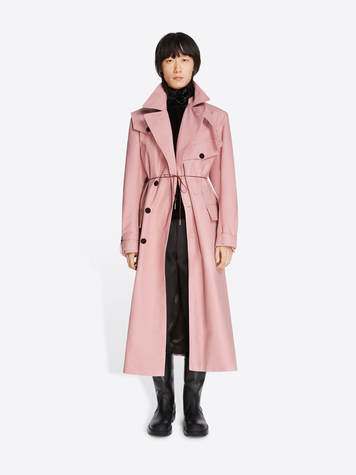 Model wearing a black turtleneck, black pants, black leather boots and a rose colored cotton trench coat on a white background